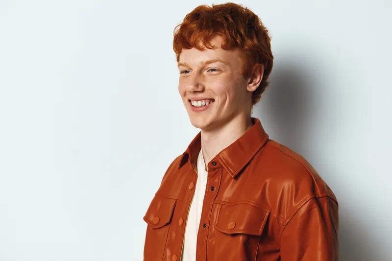 Young man with red hair smiling wearing stylish brown leather jacket and white shirt standing against white background with confident and happy expression.