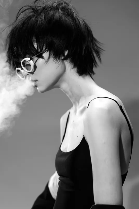 Black and white portrait of a young woman with short hair and glasses exhaling smoke, stylish and moody atmosphere.