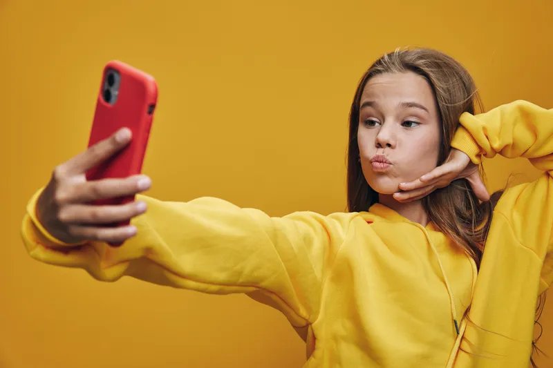 Teenage girl in yellow sweater taking selfie with smartphone on orange background. Young female posing with hand near face for social media photo content.