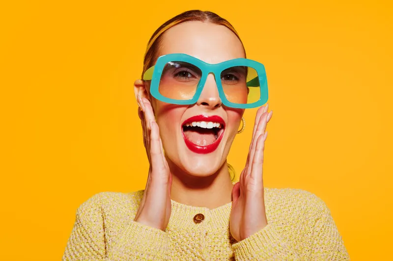 Happy woman with bright red lipstick and large turquoise glasses smiles widely, wearing a yellow sweater against an orange background. Concept of joy, fashion, and vibrant style.