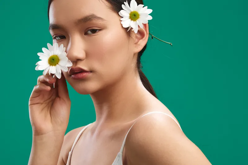 Asian woman with daisies in her hair poses against a vivid green backdrop, conveying a calm, natural beauty and contemporary elegance in a studio portrait.