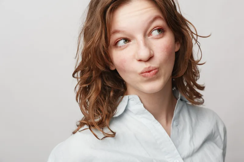 Young woman with curly hair wearing white shirt looks sideways with playful expression on light background. Concept of casual style, youth, and natural beauty.