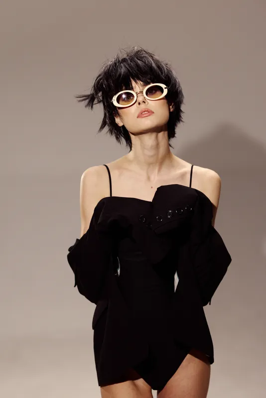 Stylish woman in black dress with sunglasses posing confidently against gray background with a modern fashion vibe.