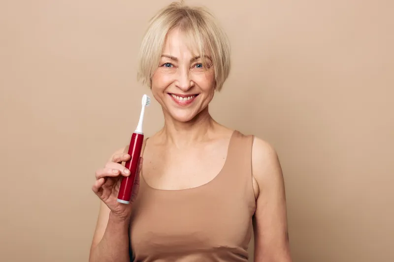 Smiling mature woman with short blonde hair holding electric toothbrush on beige background. Concept of dental care, oral hygiene, wellness, and healthy lifestyle for seniors.