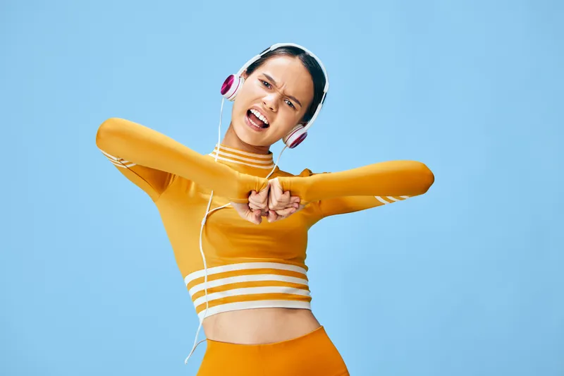 Athletic woman in an orange workout outfit dancing with headphones on a blue studio background, stretching and smiling, showcasing a vibrant fitness routine and energetic lifestyle.