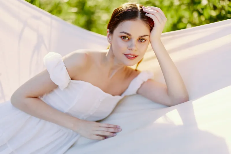 Young woman in a white dress relaxing in a hammock, exuding peaceful emotions amidst a lush green backdrop, ideal for concepts of tranquility and summer bliss