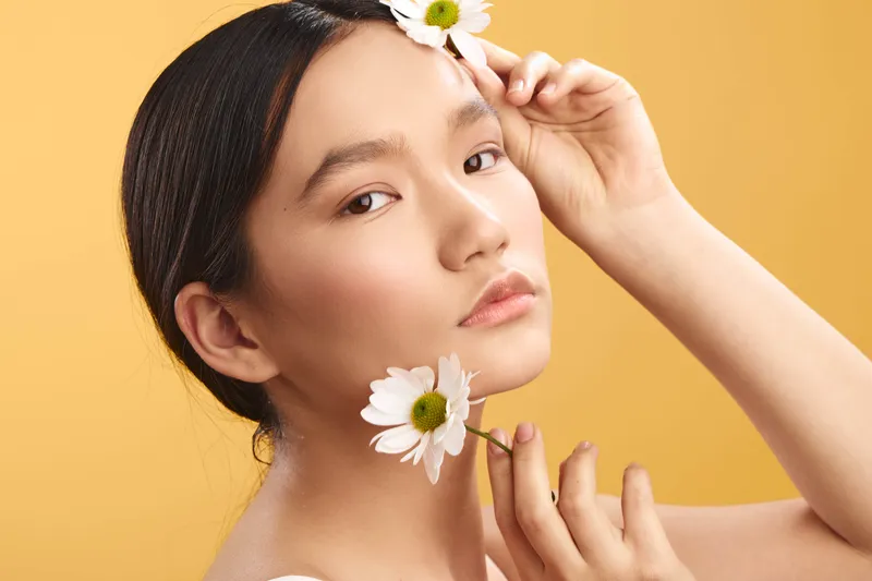 asian woman with delicate features poses among white daisies in a studio portrait, set against a warm yellow backdrop, highlighting natural beauty and a serene, refined mood.