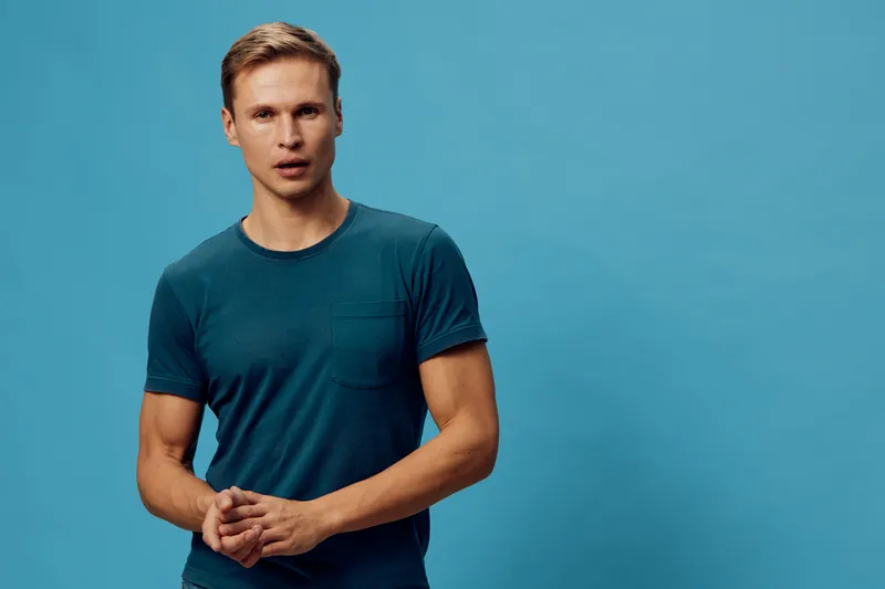 Young man with blonde hair wearing navy blue t-shirt standing against solid blue background, looking surprised and confused, isolated in studio portrait, casual clothing, expressive face, modern style