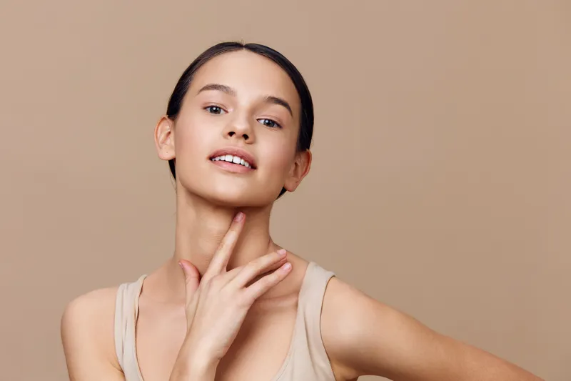 Young woman with neutral expression, wearing a beige sleeveless top, presenting natural beauty against a soft beige background for beauty and skincare concepts