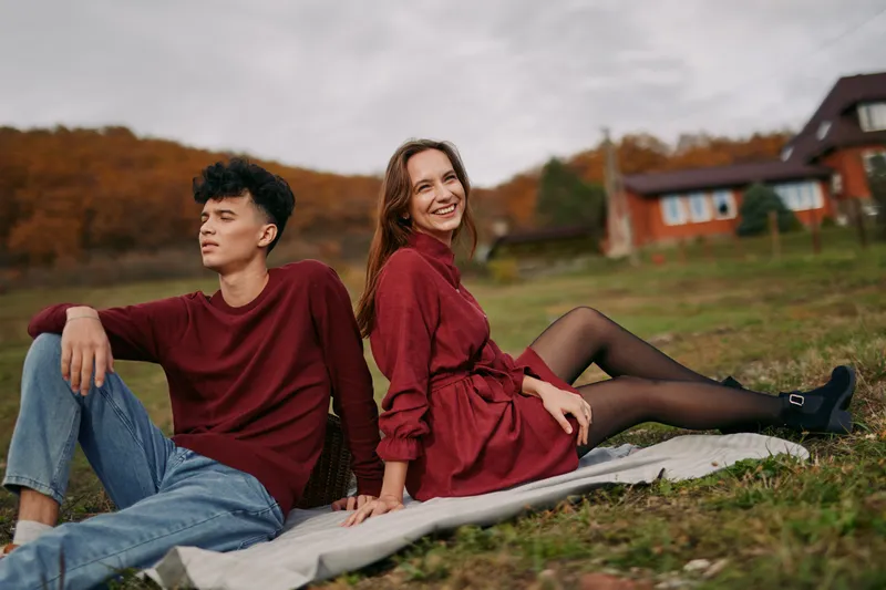 Young couple sitting on a blanket in an autumn meadow near a rustic cottage, natural poses and genuine smiles conveying authenticity and credibility in a relaxed, intimate outdoor portrait scene.