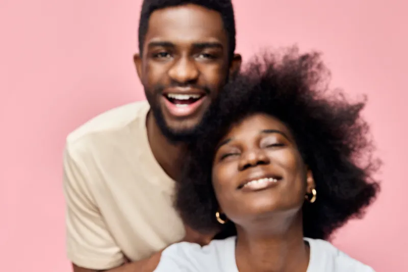 Smiling Black couple with natural hair against a pastel pink background, conveying joy and love in a playful, vibrant atmosphere, perfect for celebrating relationships