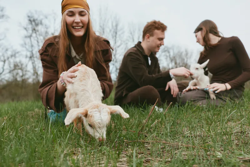 Young woman with long hair happily feeds a baby goat while two friends sit on grass in casual clothes enjoying a relaxed lifestyle moment outdoors in early spring.