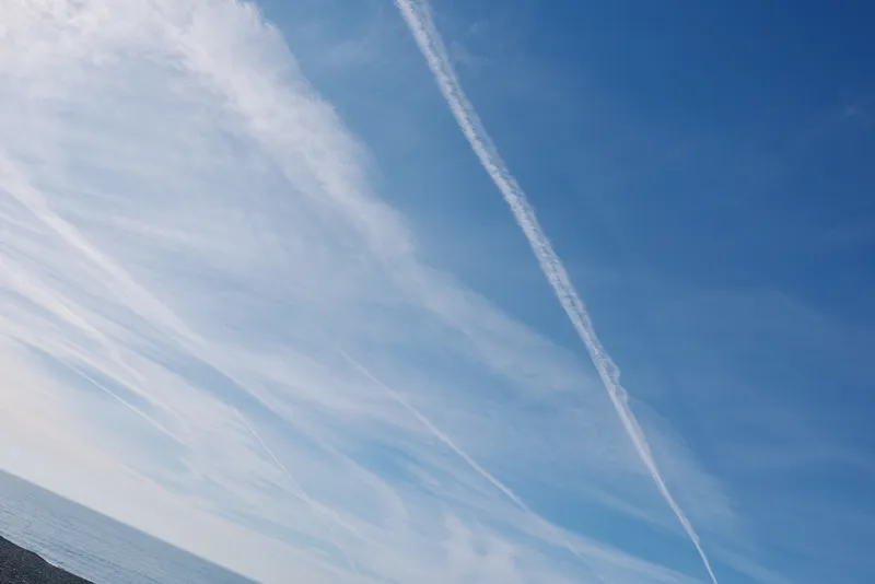 Blue sky over calm ocean with wispy clouds and diagonal contrails crossing the horizon, creating a serene seascape ideal for travel, nature, weather and inspirational stock imagery.