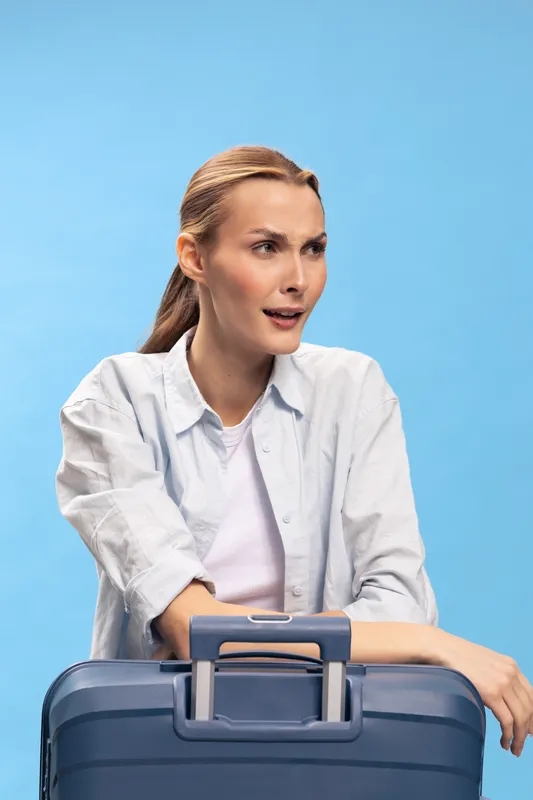 Young woman with blonde hair leans on blue suitcase and looks to the side on blue background. Concept of travel, waiting, and vacation planning with casual outfit and natural light.