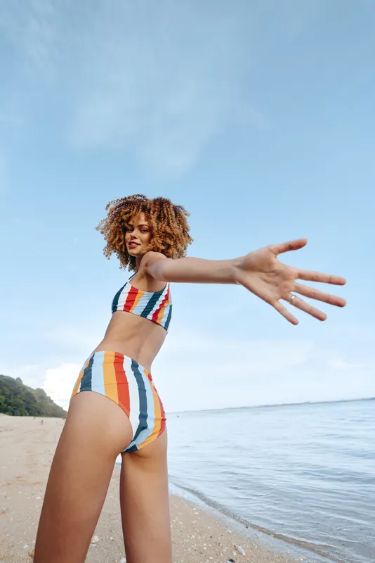Woman with curly hair in a colorful striped swimsuit, standing on a beach by the ocean, pose with arms stretched forward, blue sky, summer outdoors, vacation mood