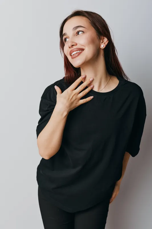 Woman smiling and looking up with hand near neck in casual black shirt standing against light background. Portrait of happy young female with natural makeup and long hair indoors.