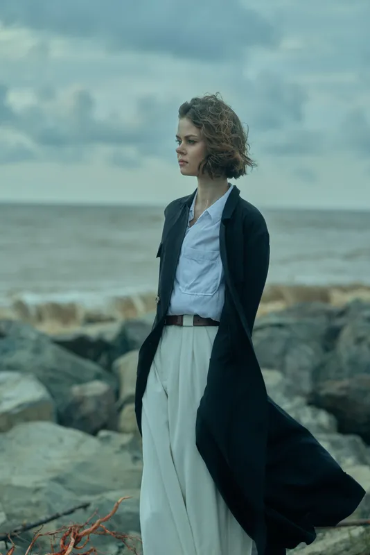 Woman in a flowing coat stands on a rocky shore, wind catching fabric, sea and cloudy sky behind for a dramatic coastal mood