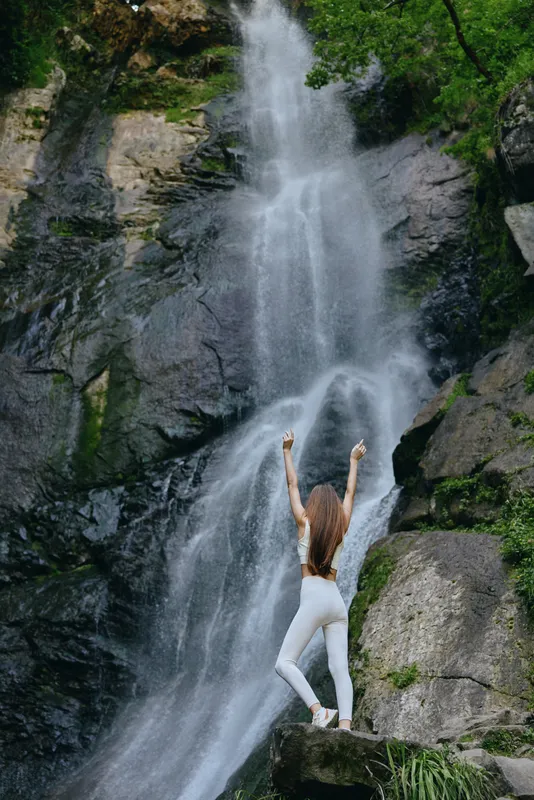 Woman in white activewear standing with arms raised near a tall natural waterfall surrounded by green foliage and rocky cliffs during daylight.