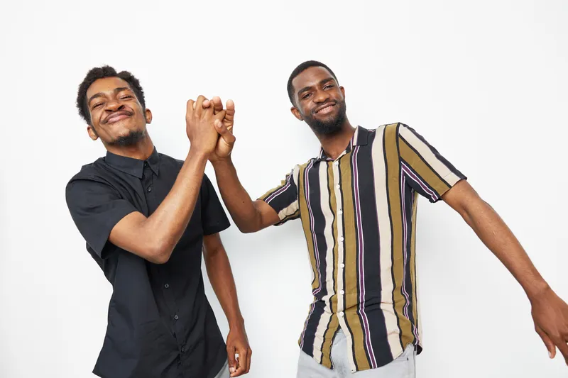Two black men share a joyful moment, smiling and celebrating together in a bright setting, showcasing friendship and camaraderie in a lively atmosphere.