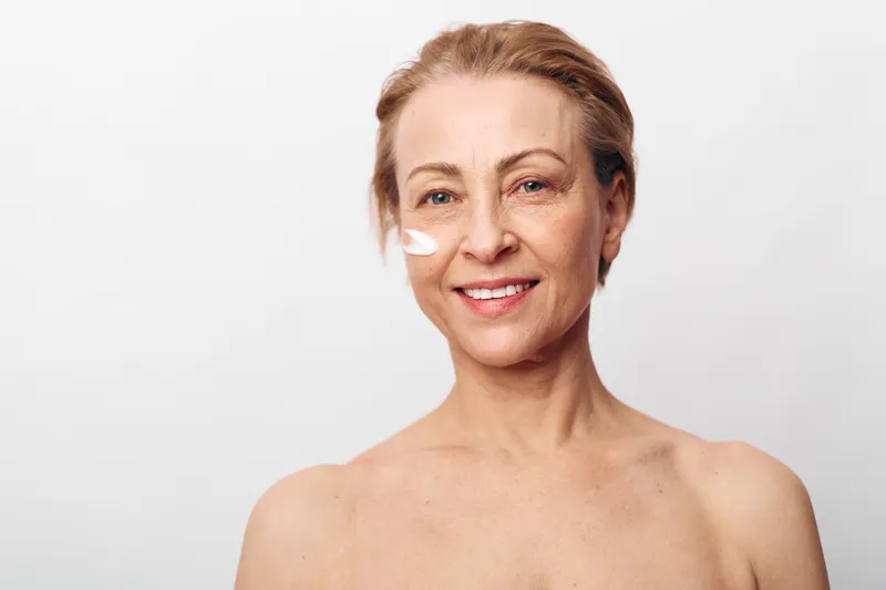 Smiling mature woman with light skin applies white face cream on cheek. Close-up portrait on plain background. Concept for skincare, beauty, aging, self-care, and natural wellness routine.