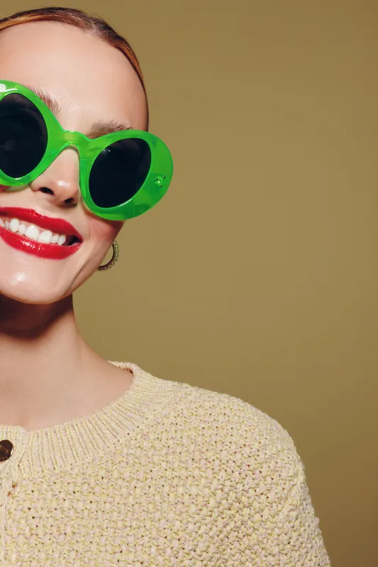 Smiling woman with bright red lipstick wears large green sunglasses and beige knitted sweater. Close-up portrait on plain background. Concept of fashion, style, and confident mood.