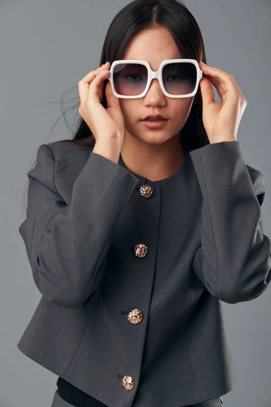 Young woman in grey jacket adjusting large white sunglasses with both hands, studio portrait on grey background showcasing fashion, style, beauty, and confident look in casual outfit.