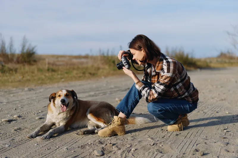 Outdoor photography scene with a woman crouching on a sandy beach and a calm dog beside her as they document nature and moments during a casual hobby session