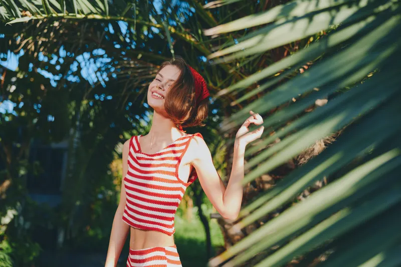 A cheerful woman in a striped red and white crop top smiles brightly while enjoying a sunny day among lush palm leaves, creating a vibrant, carefree outdoor mood and lively atmosphere.