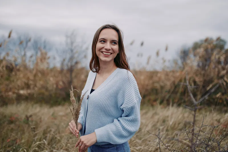 Smiling young woman in a cozy sweater holds wild grass in a windswept field, captured with natural light, relaxed posture and a genuine smile to convey authenticity and honest everyday life.