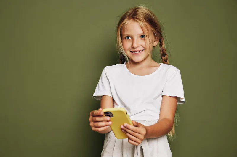 Smiling young girl in white shirt holding yellow smartphone looking at camera. Studio portrait on green background. Concept of childhood, technology, communication, and happiness.