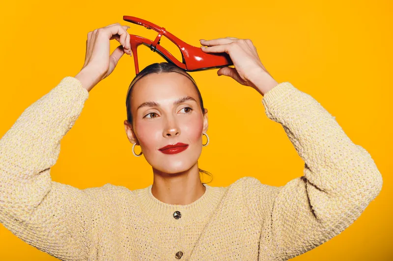 Young woman in beige sweater holds red high heel shoe above head against bright yellow background. Concept of fashion, style, creativity, and playful mood in studio lighting.
