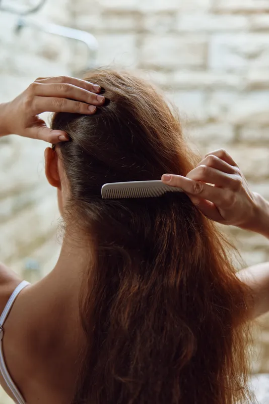 woman combing long brown hair from behind, natural light, casual white top, textured background, hair care routine, gentle grooming moment