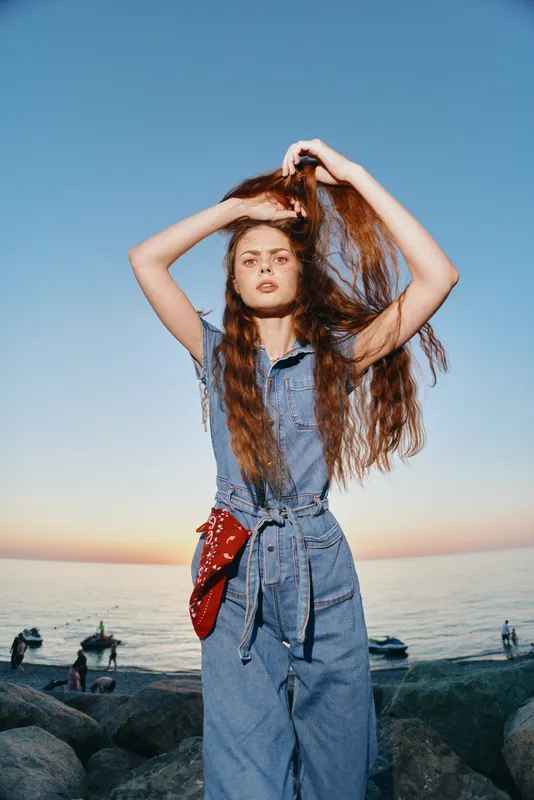 young woman standing by the sea at sunset wearing denim outfit and red belt bag with long hair flowing in the wind under clear blue sky