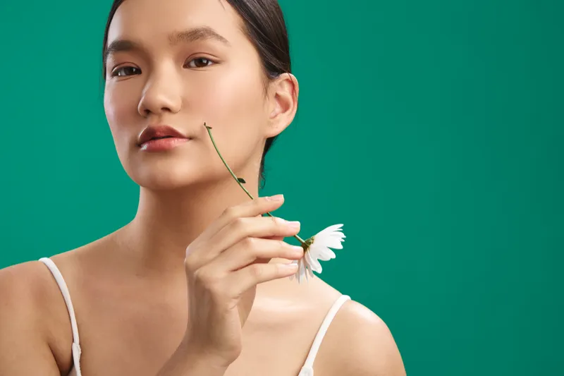 Asian woman with calm poise is posed against a vivid green backdrop, holding a delicate white flower near her lips as soft light highlights her features.