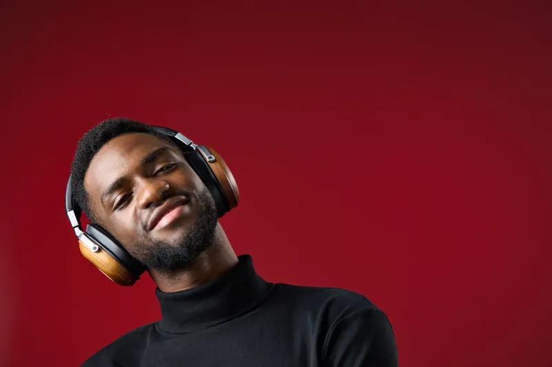 Stylish young Black man enjoying music with headphones against a vibrant red background, exuding confidence and relaxation