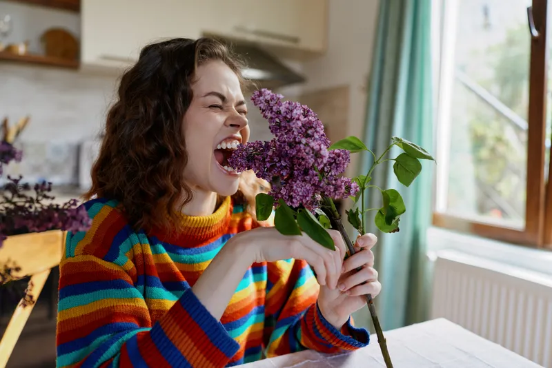 Young woman with curly hair sits at a sunlit kitchen table, biting a purple lilac bouquet, while wearing a colorful striped sweater, conveying playful, cozy home mood and self care.