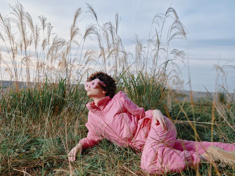 Outdoor portrait of a woman in a pink jumpsuit reclining in tall grass under a clear sky, capturing a relaxed, carefree moment in nature and countryside breeze with soft lighting.