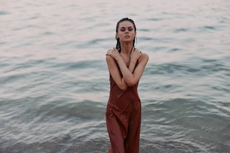 A lone woman stands in shallow sea water wearing a rust brown dress, arms crossed, calm gaze, and a soft light that creates a minimalist coastal mood.