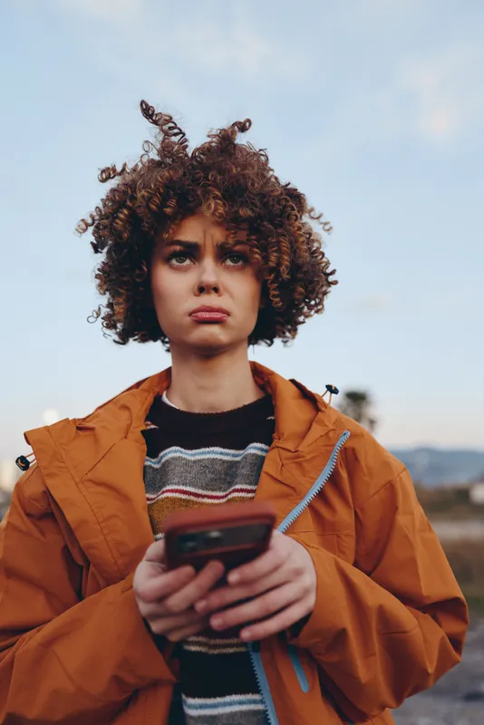 Woman lifestyle outdoors portrait, curly hair woman in orange jacket holds smartphone, looking alert at blue sky, casual street style, natural light, upper body, young adult, travel mood