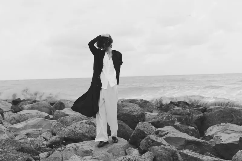 Man on a rocky shore wearing a long coat and light trousers, wind blowing the garment as waves crash nearby in a moody black and white coastal scene