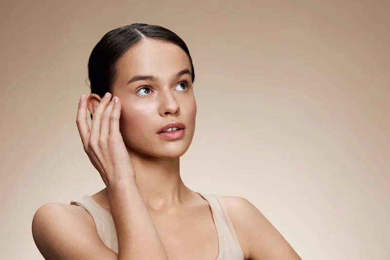 Young woman with natural makeup, gently touching her face, showcasing healthy skin and a calm expression against a neutral beige background