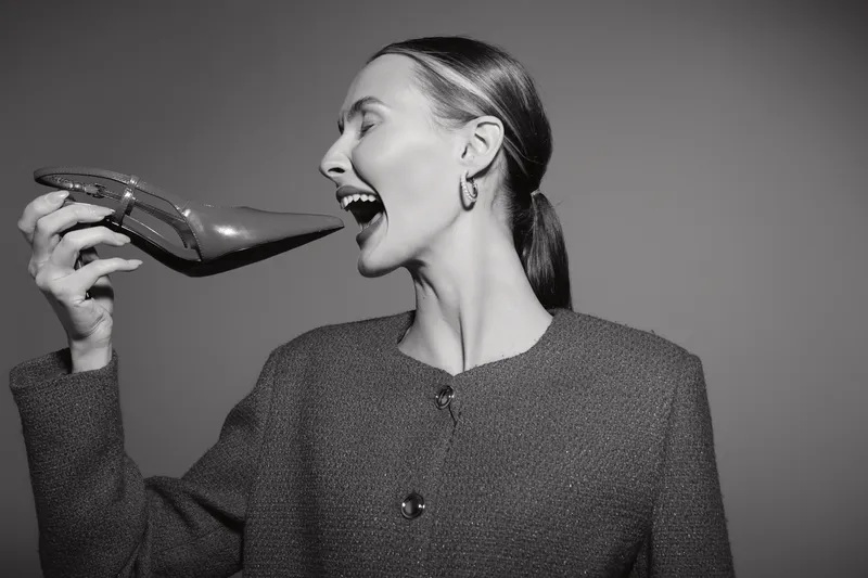A smiling caucasian woman with light skin holds a shiny high heel shoe near her face in monochrome. Concept for fashion, style, and joyful attitude in studio lighting.