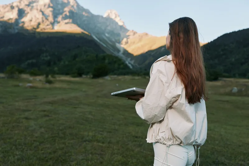 A woman stands in a wide alpine meadow, holding a notebook and looking toward distant peaks. She wears light outdoor gear, conveying focus, exploration, and calm productivity.