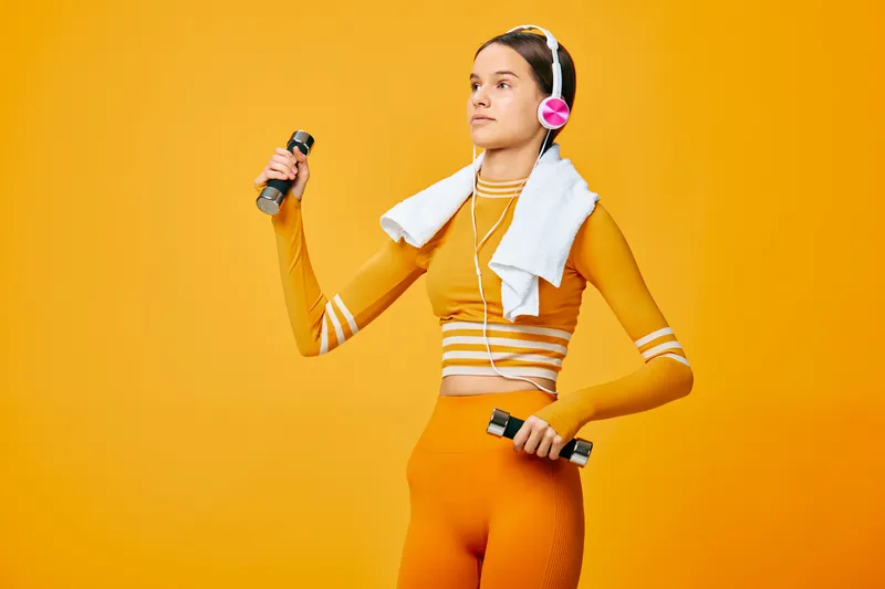 Fitness woman in orange athletic wear lifts dumbbells with a towel over her shoulders, wearing headphones, ready for a dynamic workout in a bright studio.