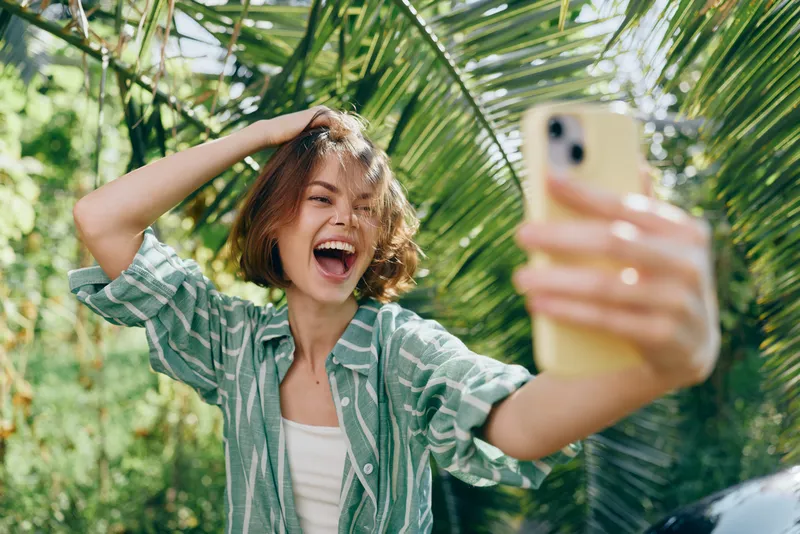 Cheerful woman takes a sunny selfie outdoors, laughing with carefree energy and a relaxed pose under lush foliage.
