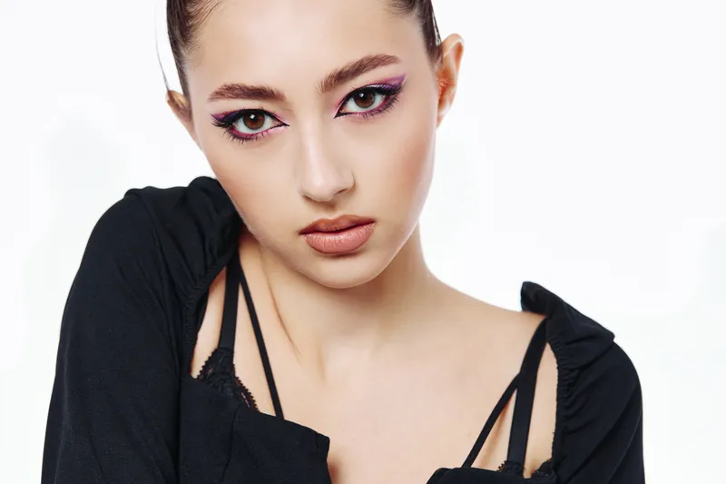 Young woman with creative vibrant makeup and eyeliner poses wearing a stylish black top. Close-up on a clean white background, highlighting beauty and fashion concepts.