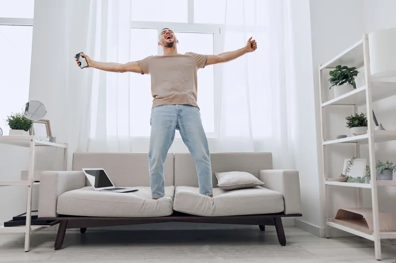 Energetic man celebrating at home, expressing joy with outstretched arms. Bright, modern living room with natural light and minimalistic decor creates a refreshing atmosphere.
