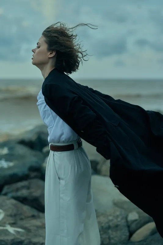 Wind cloak by the beach, a woman in a white dress stands by the ocean, windblown fabric fluttering, capturing movement, coastal elegance and a moody maritime atmosphere