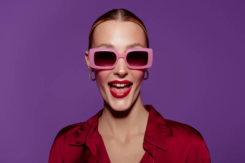 Happy young woman with pink sunglasses and red lipstick smiles widely against purple background. Fashion portrait with bright colors and joyful expression. Concept of style and confidence.