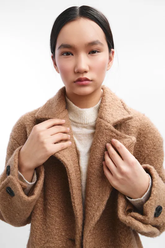 Asian woman wearing a warm coat stands against a plain background, presenting a poised, contemporary portrait suitable for fashion and lifestyle contexts.